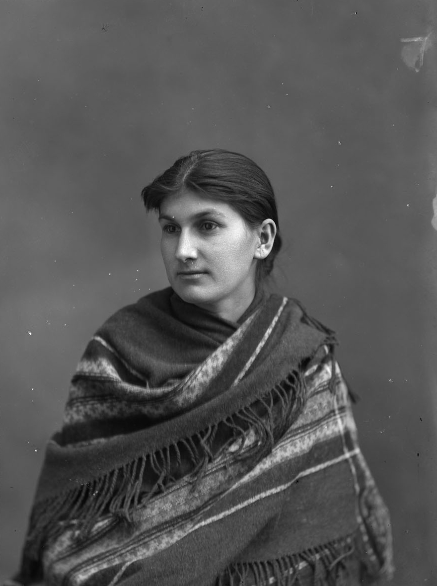 Black and white studio portrait photograph of a seated woman wearing a striped, wool shawl with fringe.